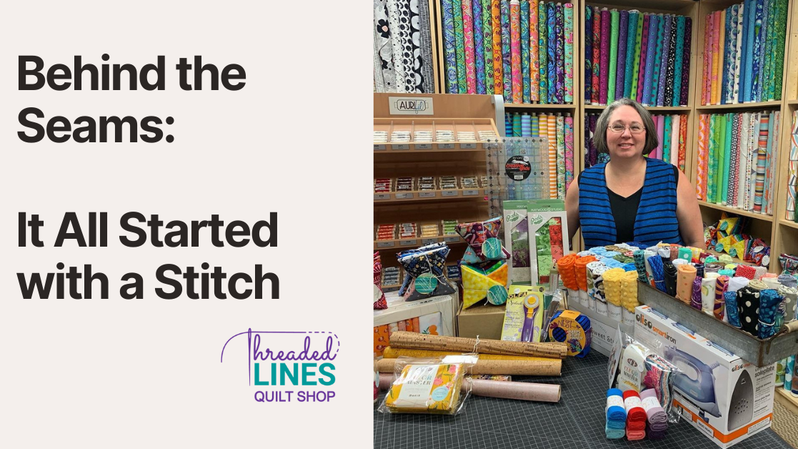 Jen standing in the original Threaded Lines Quilt Shop, smiling behind a cutting table filled with fabric, notionsa nd quilting tools. Bolts of bright, modern fabric line the walls behind her.