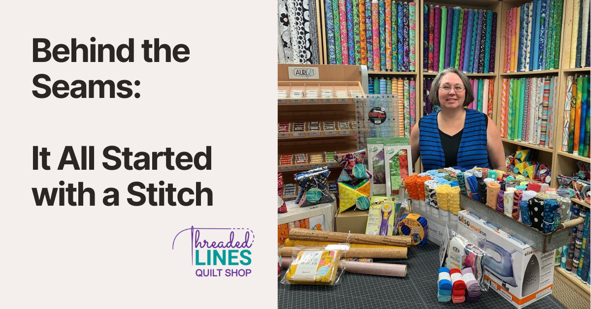 Jen standing in the original Threaded Lines Quilt Shop, smiling behind a cutting table filled with fabric, notionsa nd quilting tools.  Bolts of bright, modern fabric line the walls behind her.